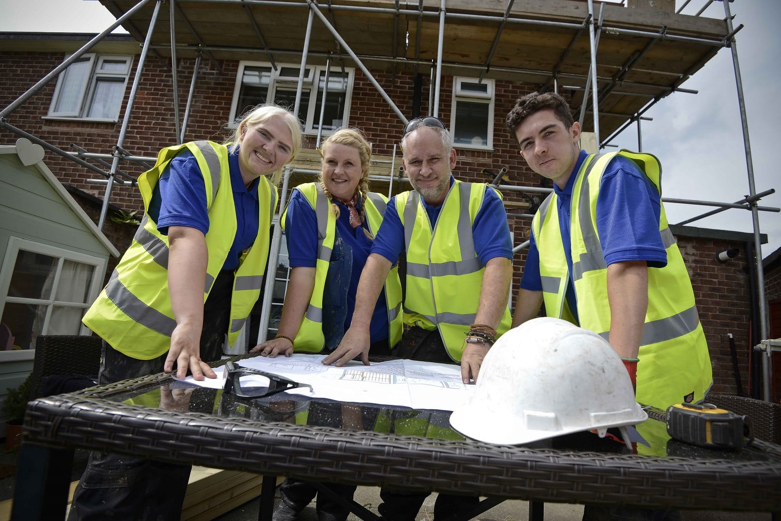 Two employers with their two apprentices, in front of a house with scaffolding with them gathered round looking at plans for the site