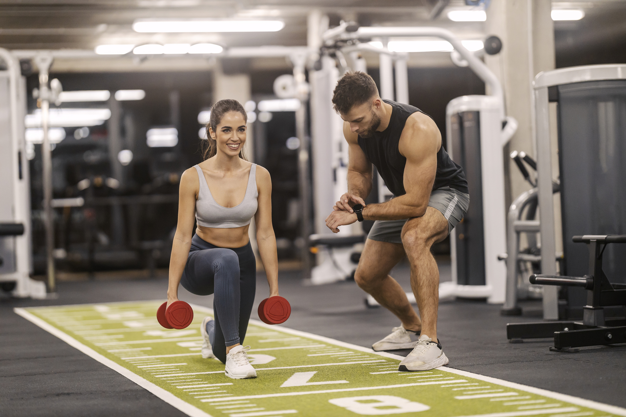 Strong fit sportswoman doing lunges with dumbbells at gym with her trainer.